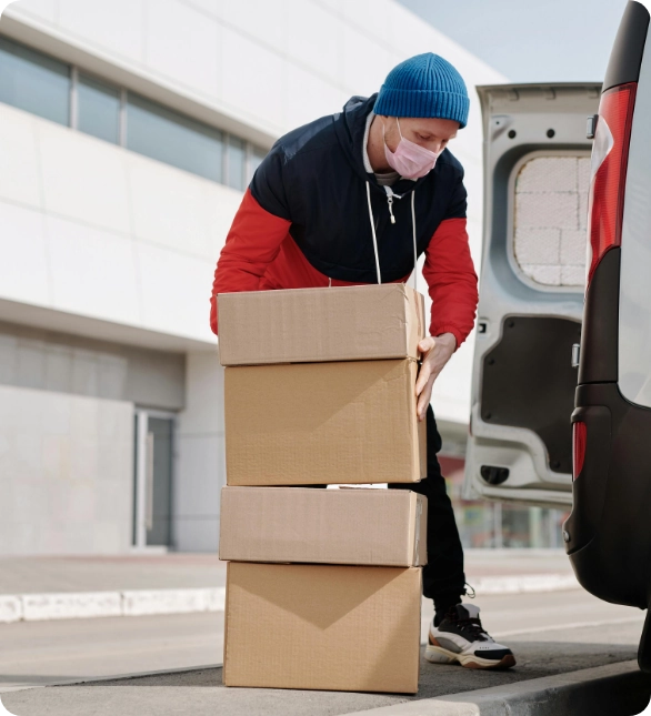 A masked man is carrying boxes beside a van, preparing for loading or unloading tasks.